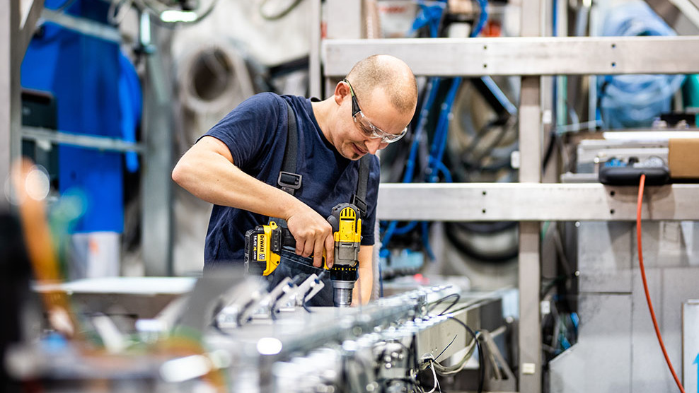 Stellenangebote Ein Monteur schraubt mit einem Akkuschrauber Teile an einer Maschine fest.